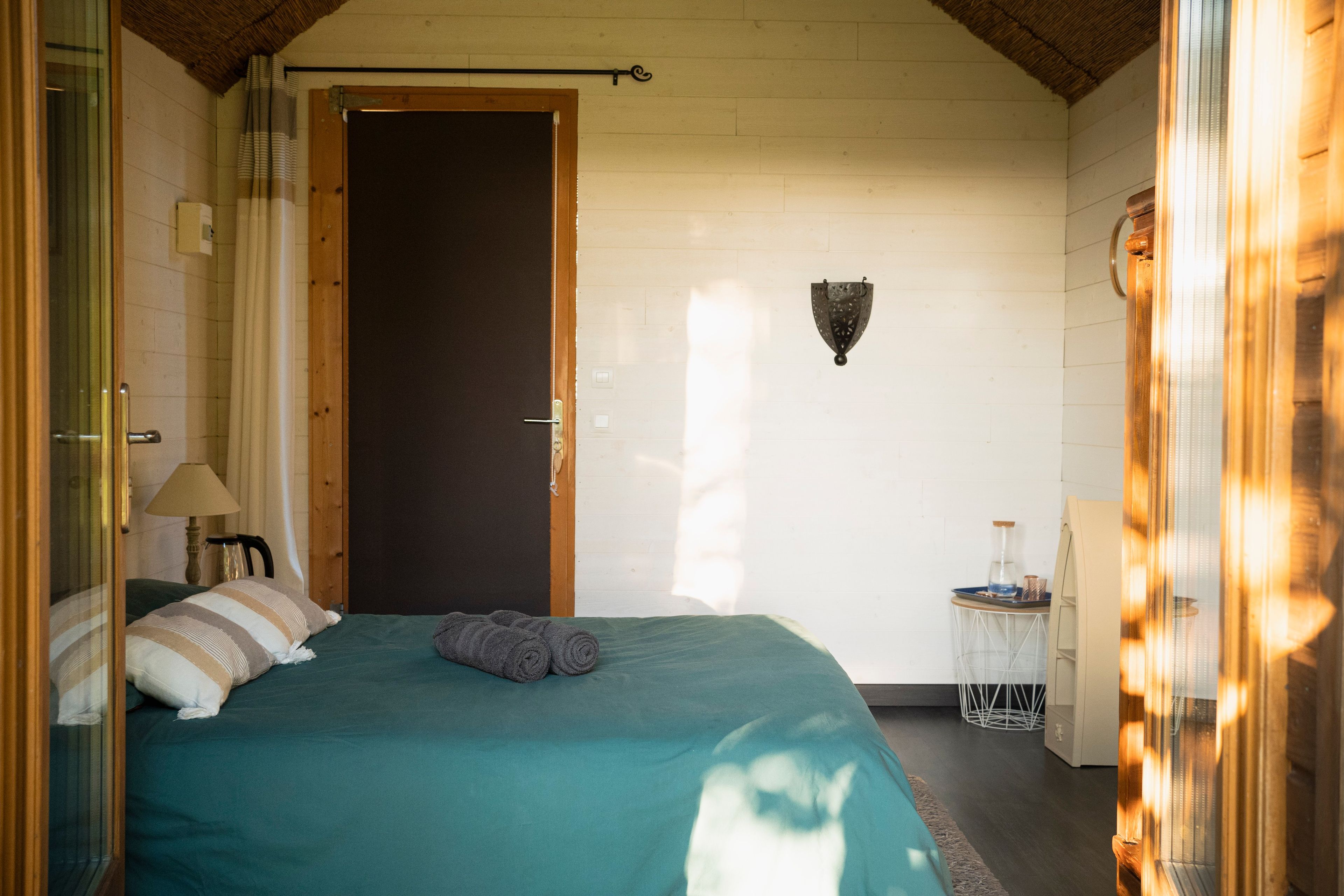 A room with a bed, a door, a wall-mounted light fixture, and a small table with a glass container.