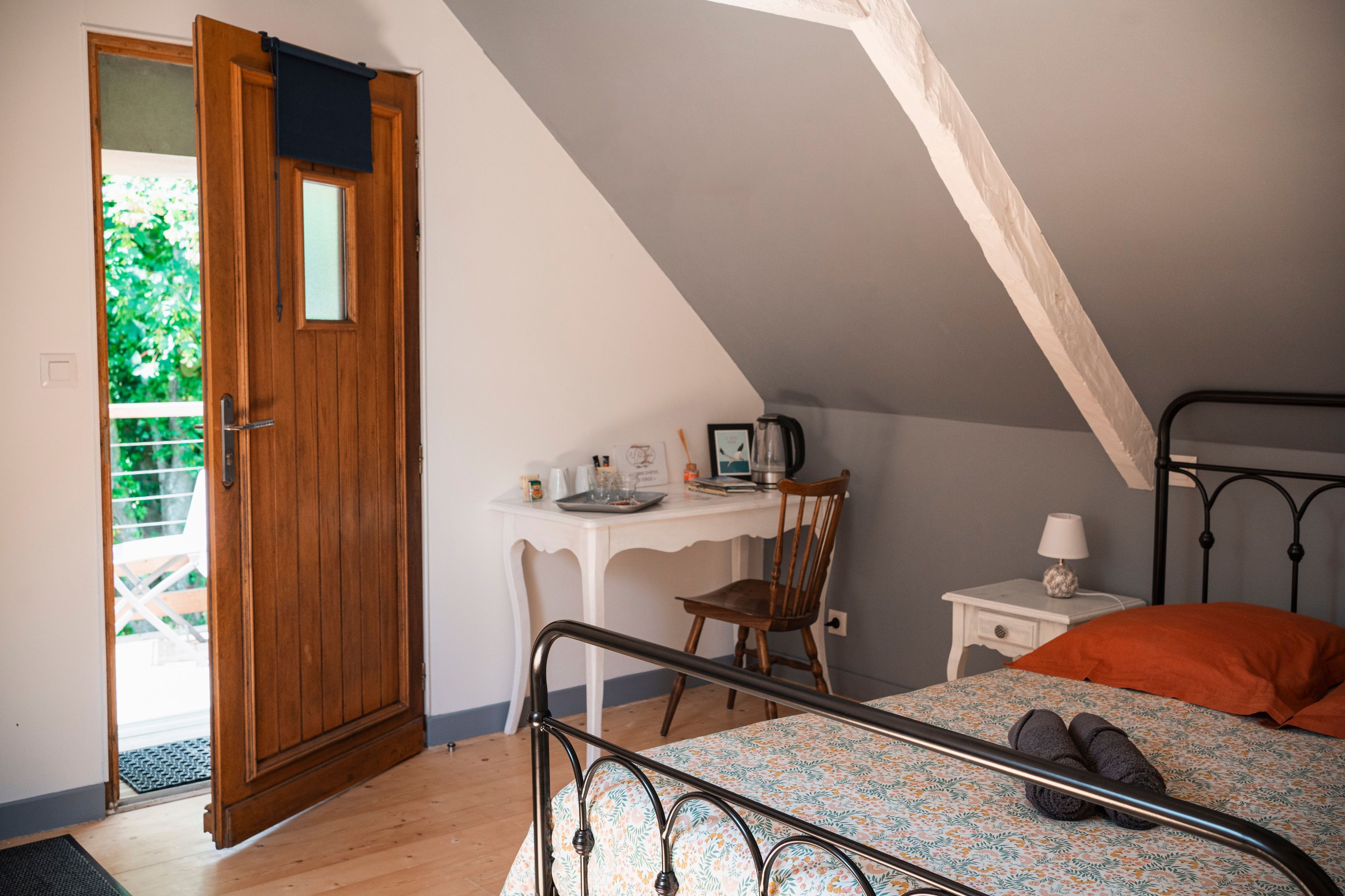 Une chambre avec un plafond en pente, une porte en bois, un bureau avec une chaise, un lit avec un cadre en métal et une table de chevet avec une lampe.
