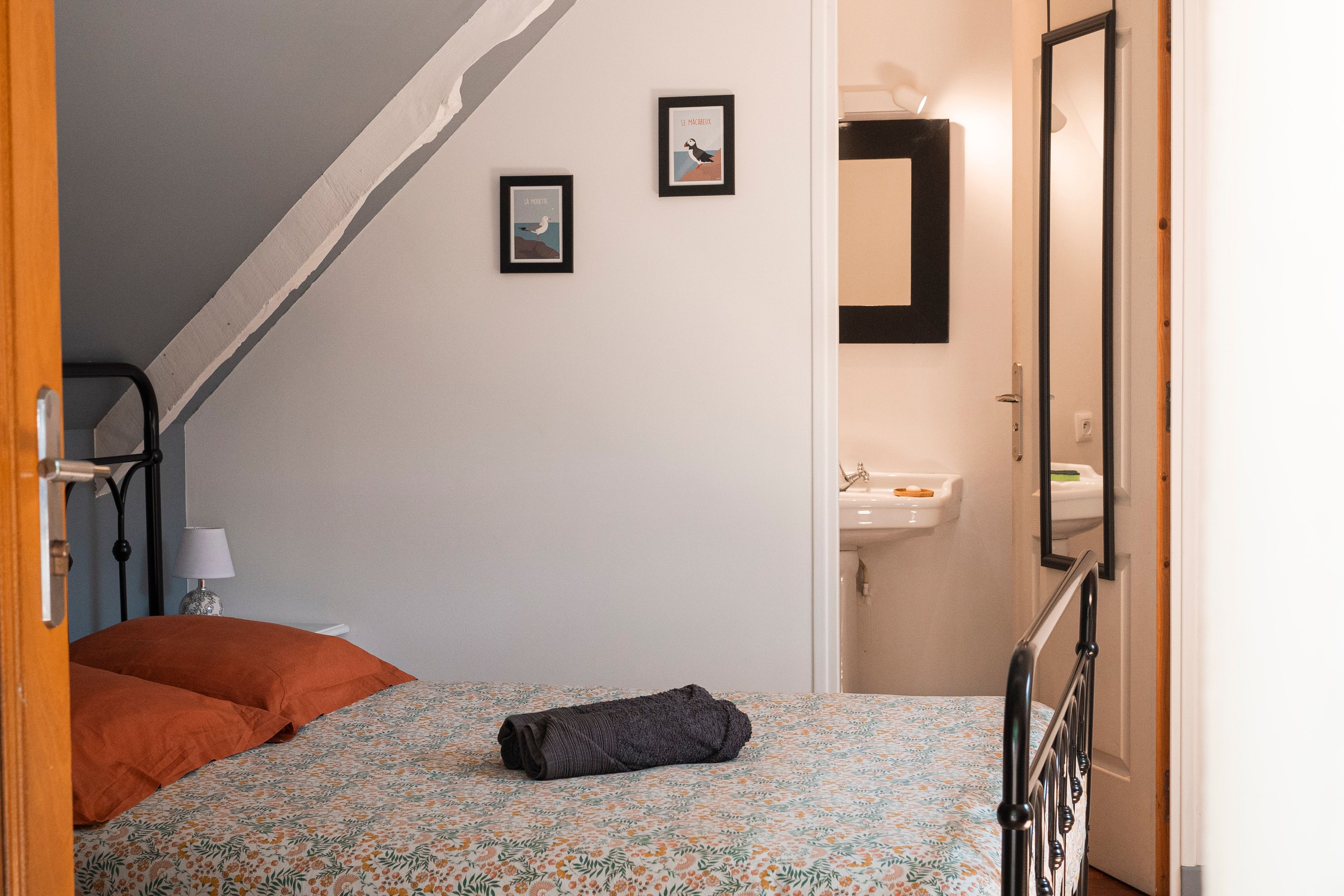 Ein Schlafzimmer mit einem Bett, einem zusammengerollten Handtuch, einem Nachttisch mit Lampe und einem an der Wand hängenden Bild.