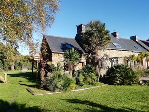 A stone house with a sloped roof, a small garden with palm trees, and a wooden deck. - Guest Room 'La Corzic' (Pléguien)