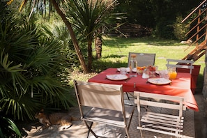 An outdoor dining area with a red table set for two, surrounded by greenery and a cat lying on the ground. - Guest Room 'La Kerguidoué' (Pléguien)