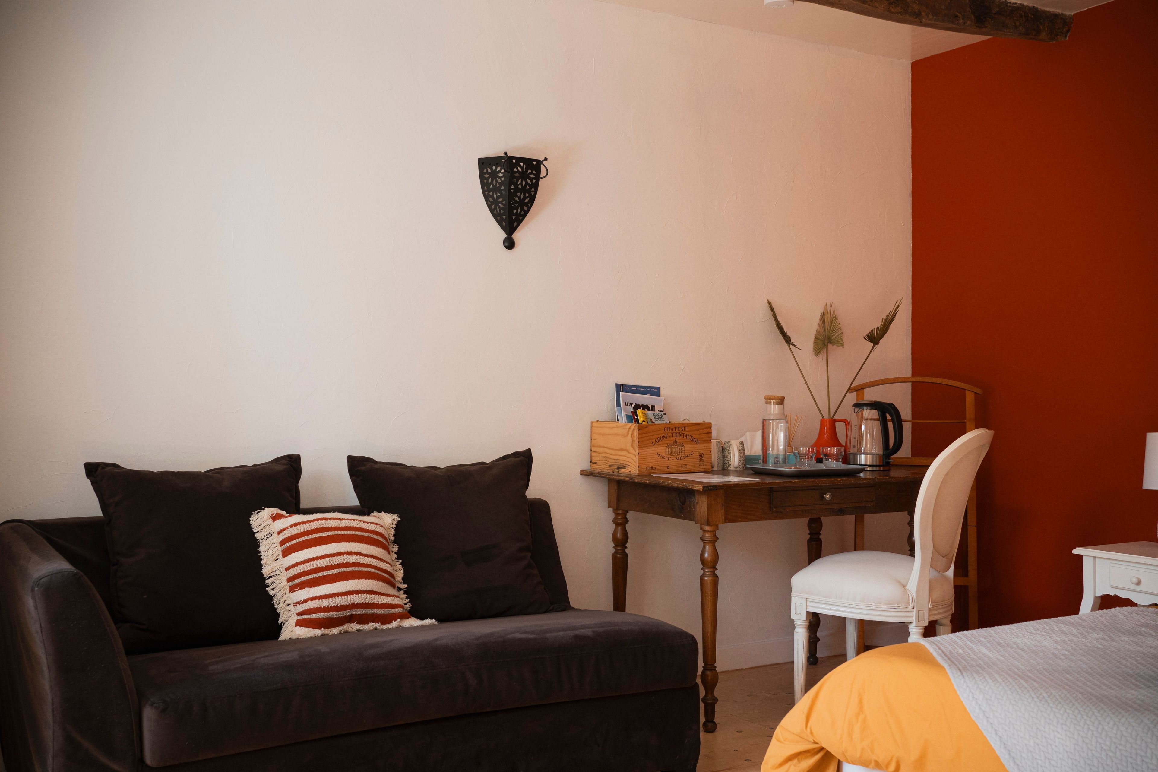 Une pièce avec un canapé de couleur sombre, un bureau en bois avec une chaise, et une petite table sur laquelle se trouvent une bouilloire et d’autres objets.