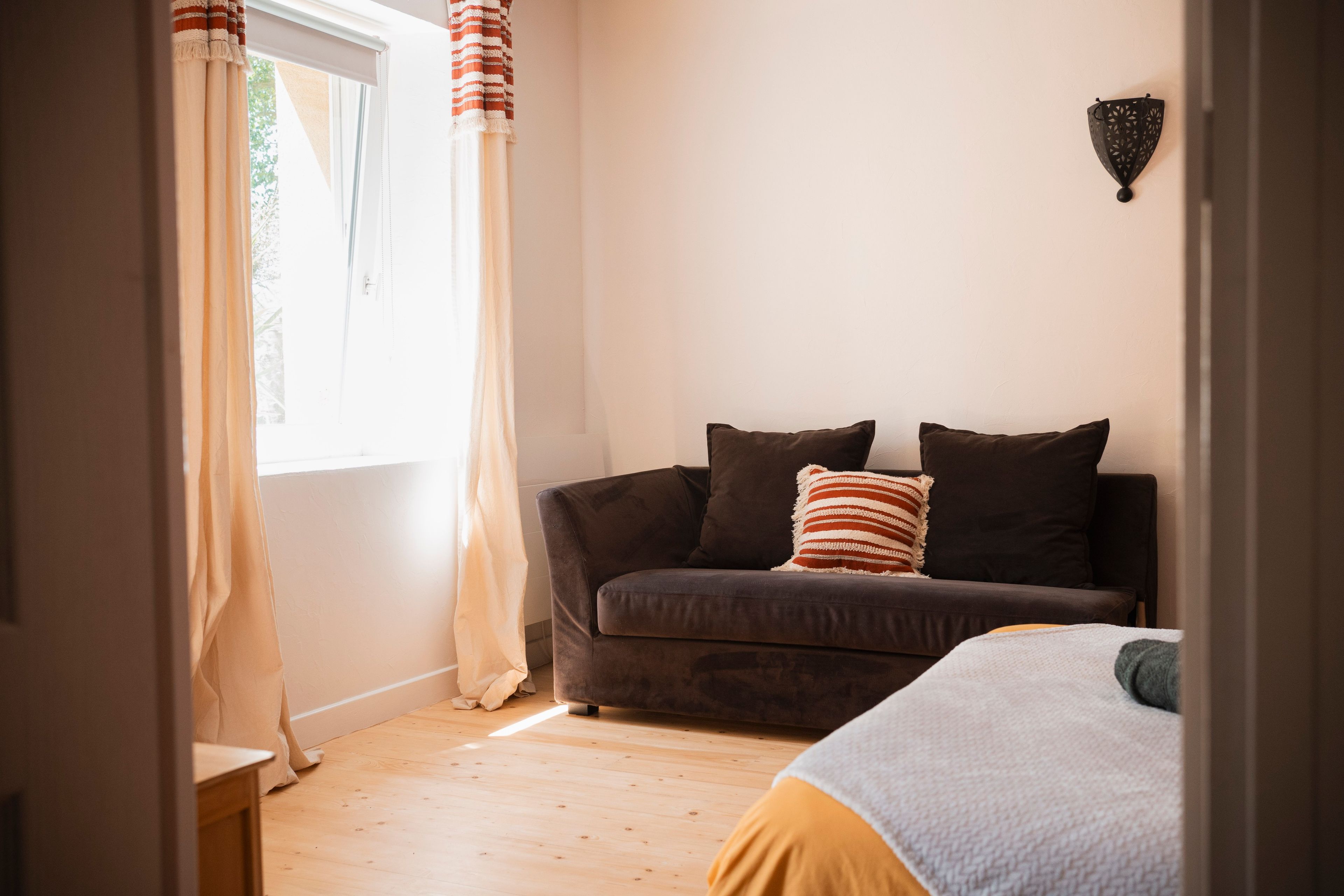 Une pièce comprenant un canapé, deux coussins, un luminaire fixé au mur et une fenêtre avec des rideaux.