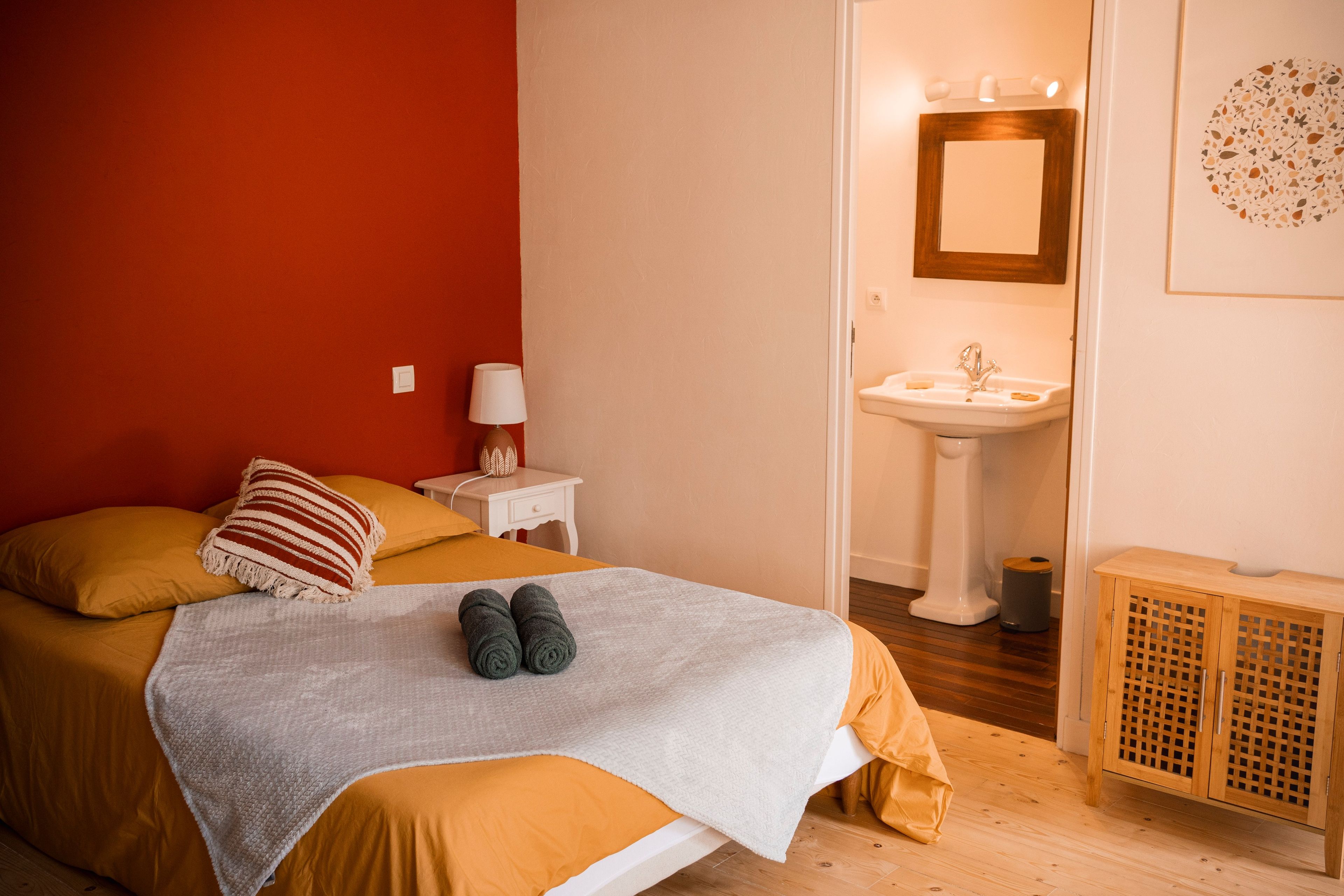 A bedroom with a bed, a bedside table with a lamp, a bathroom with a sink and mirror, and a wooden cabinet.
