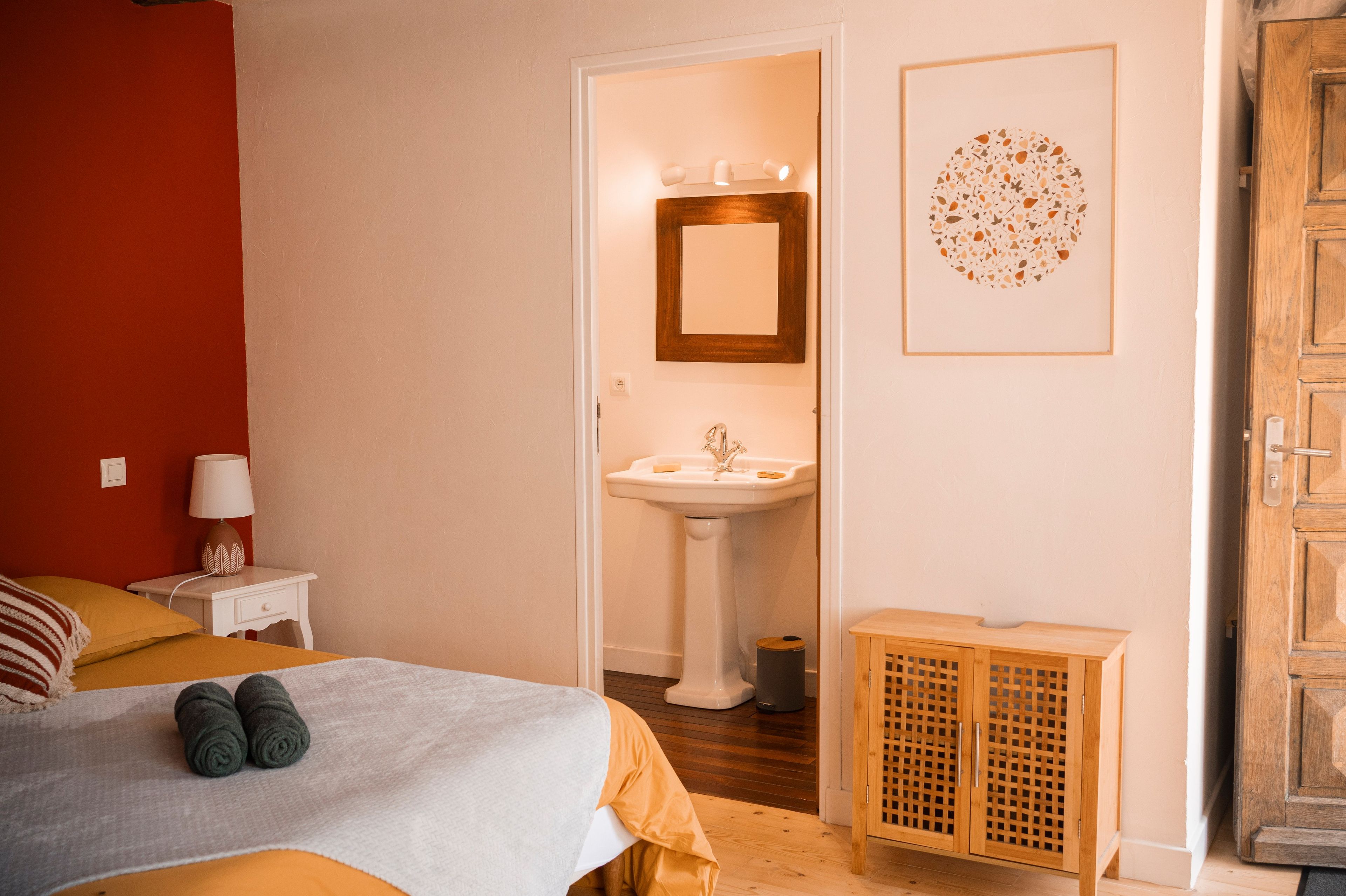 Een slaapkamer met een bed, een nachtkastje met een lamp, een houten kast, en een badkamer met een wastafel en spiegel.