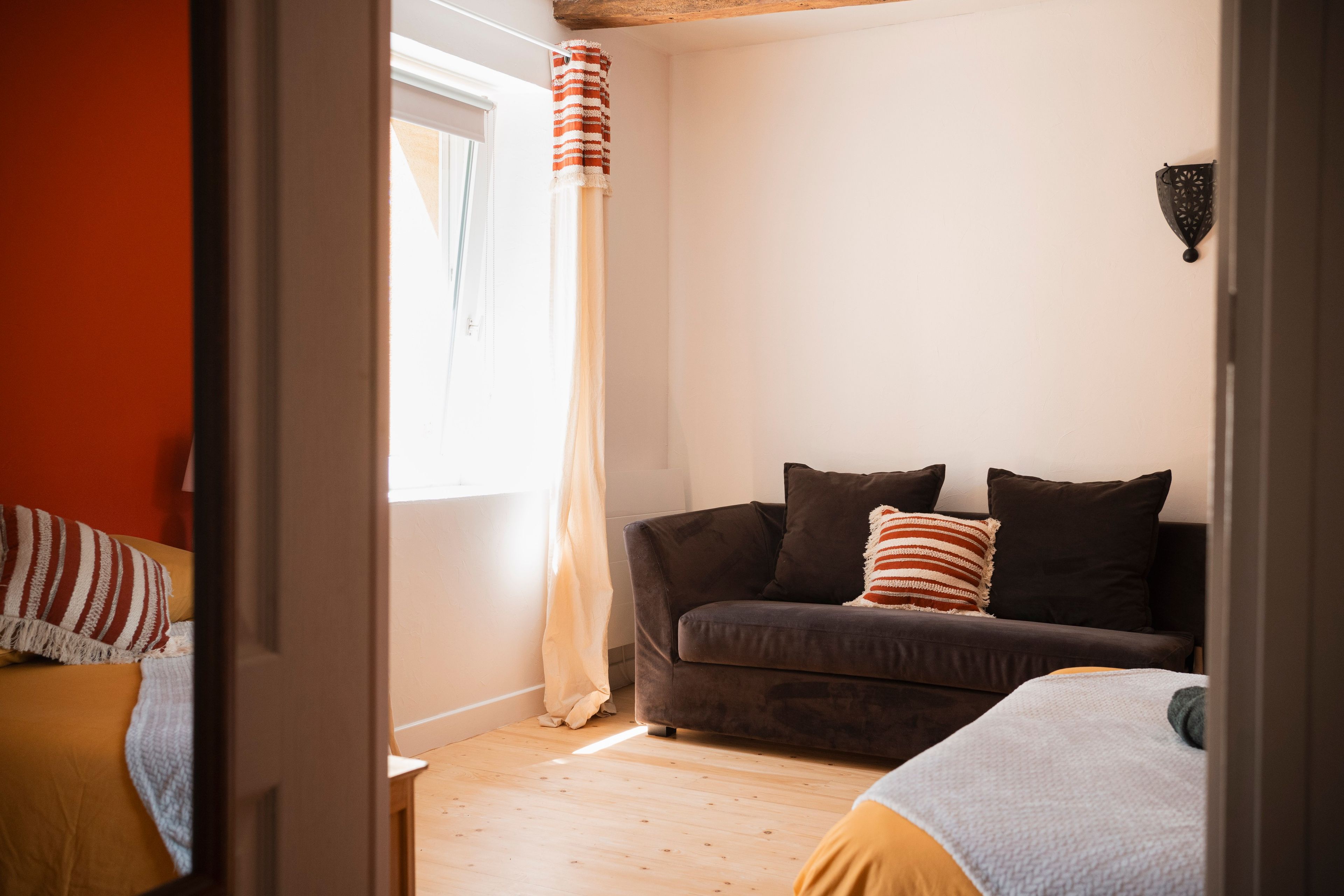 Een kamer met een houten vloer, een bank met kussens, een raam met gordijnen en een wandlamp.