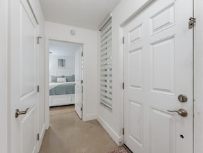 A narrow hallway with white doors, a bed with a blue bedspread, and a window with blinds. - Madeira Vista 306 (Madeira Beach)