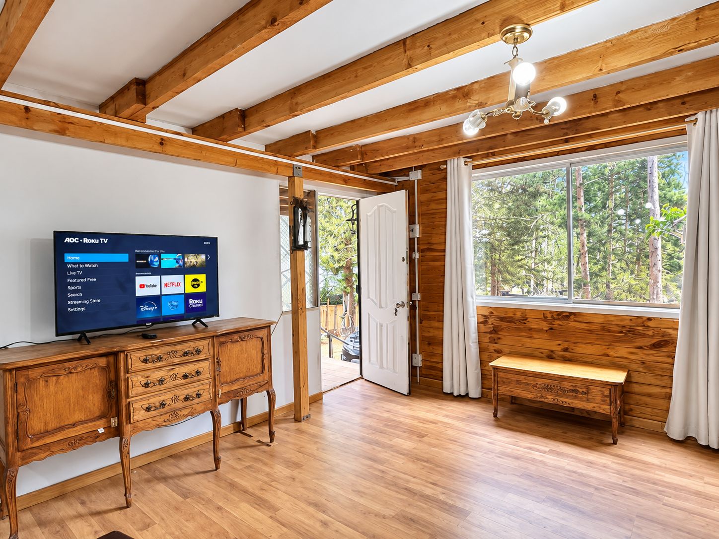 Une pièce avec un plafond en bois, un téléviseur affichant l’interface Roku, un banc en bois et une grande fenêtre donnant sur la forêt.