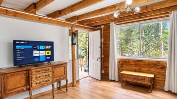 Une pièce avec un plafond en bois, un téléviseur affichant l’interface Roku, un banc en bois et une grande fenêtre donnant sur la forêt.