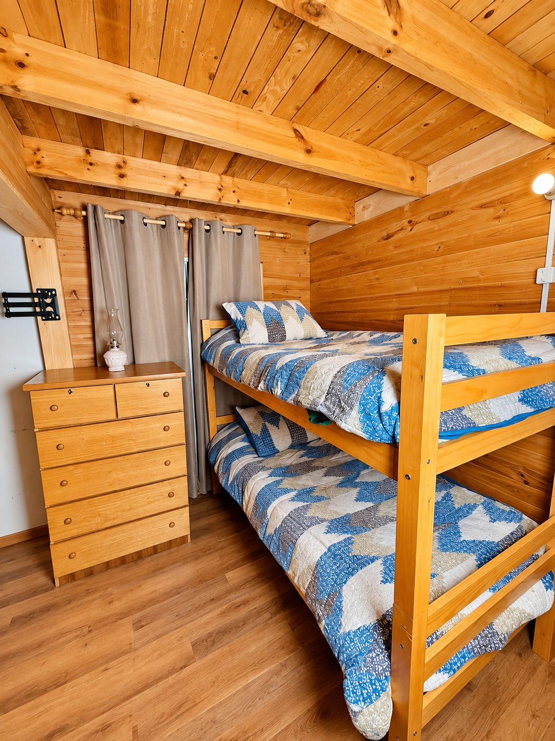 Una cama litera con una colcha estampada, un aparador de madera con cajones y una lámpara sobre un suelo de madera.