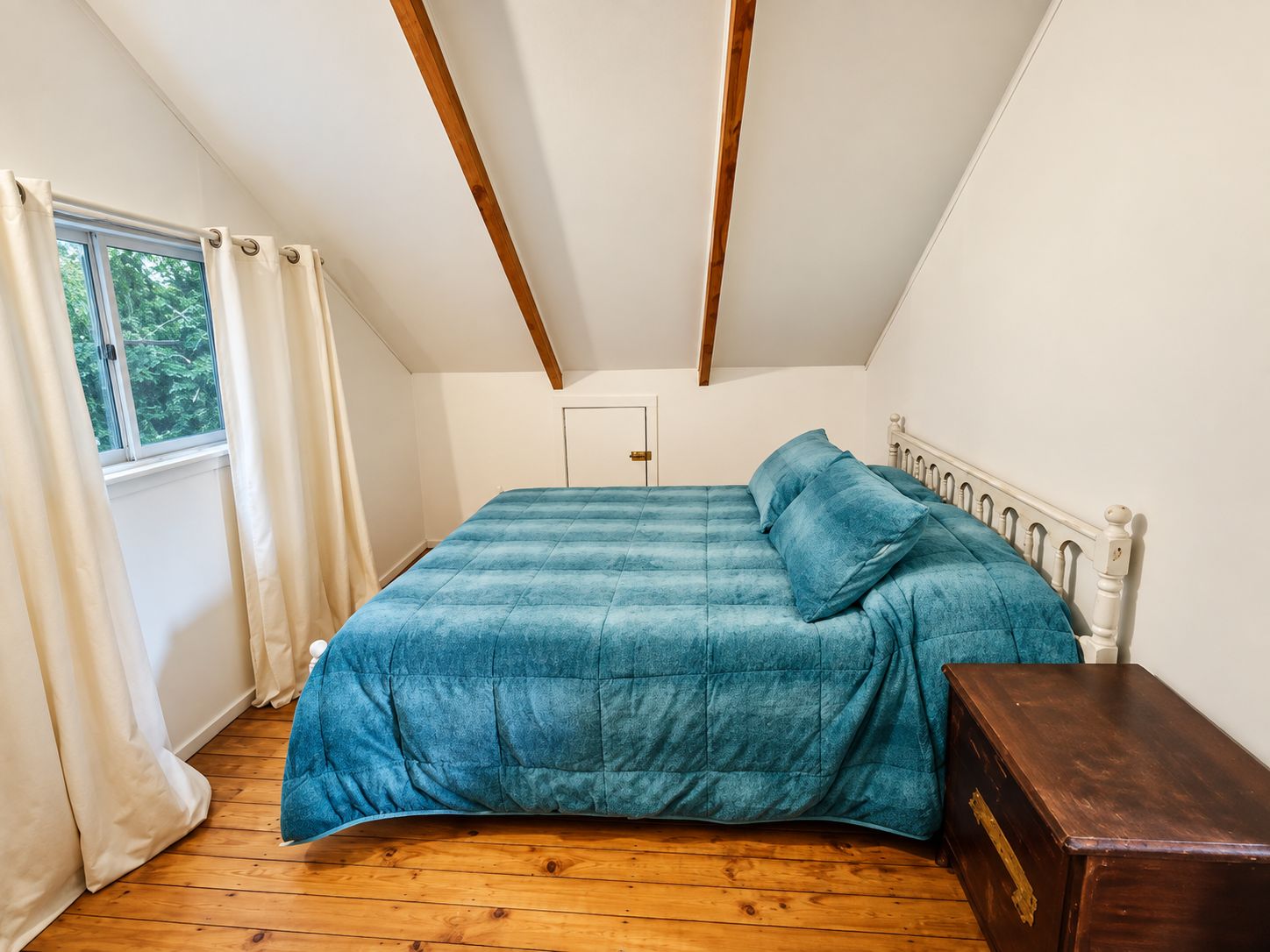 Un dormitorio con cama, piso de madera, ventana con cortinas y una puerta.