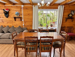 A wooden interior with a dining table surrounded by chairs, a sofa with cushions, and a window with curtains. - Cabaña del Bosque - Laguna Verde (Valparaiso)