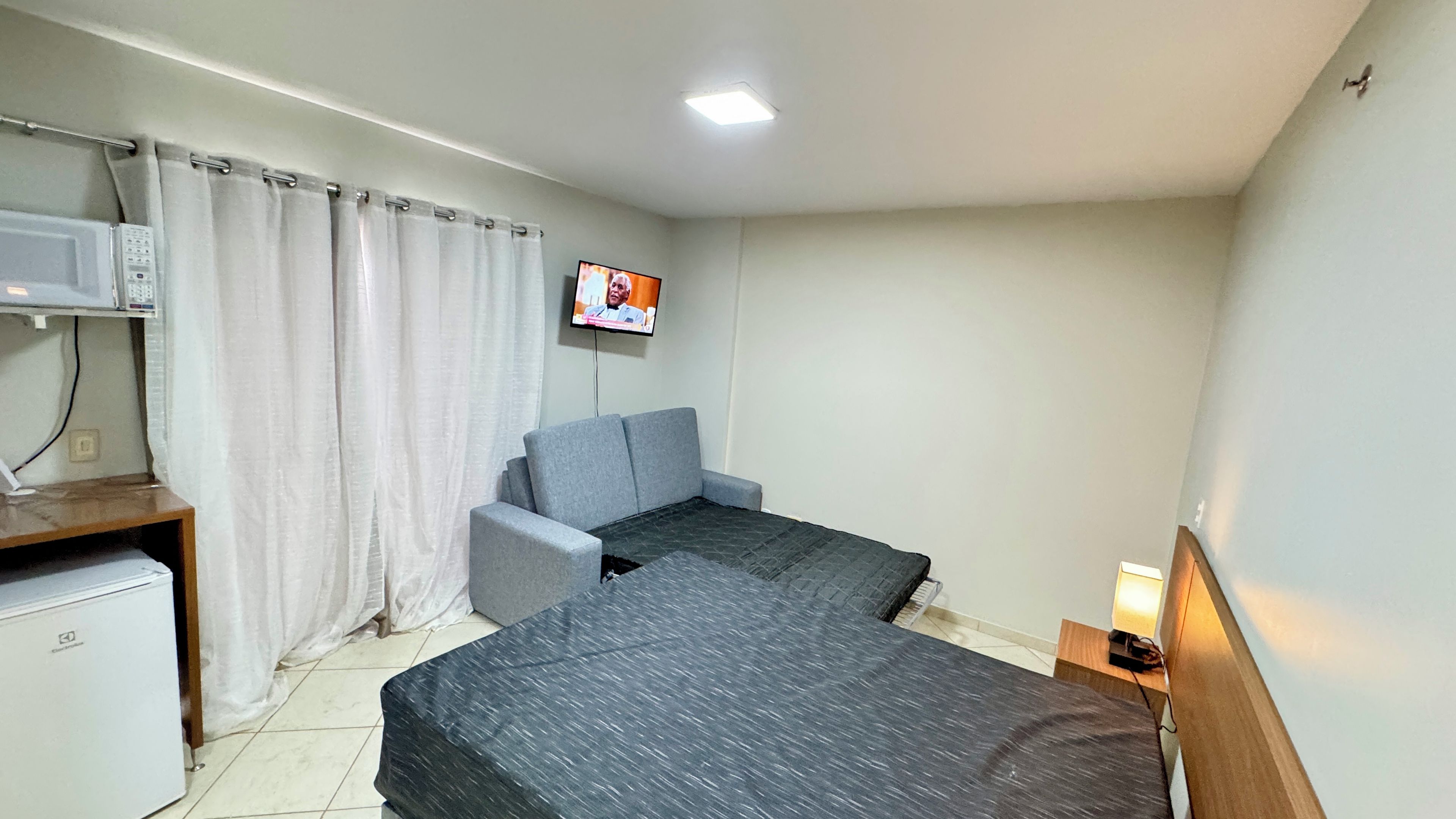 Quarto pequeno e aconchegante, com cama, escrivaninha, cadeira, televisão e mesa de cabeceira com abajur.