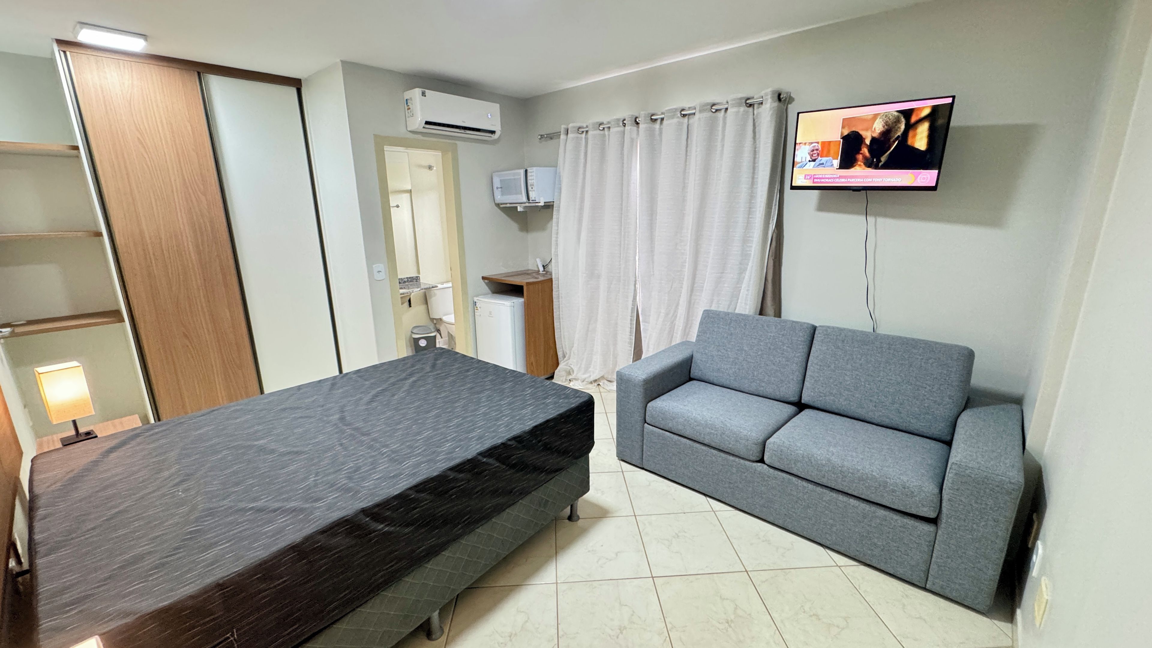 Quarto com cama, sofá cinzento, televisão e ar condicionado, além de um pequeno armário.