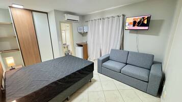 Quarto com cama, sofá cinzento, televisão e ar condicionado, além de um pequeno armário.