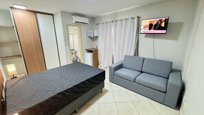 Una habitación con una cama, un sofá gris, un televisor, un aire acondicionado y un mueble pequeño.