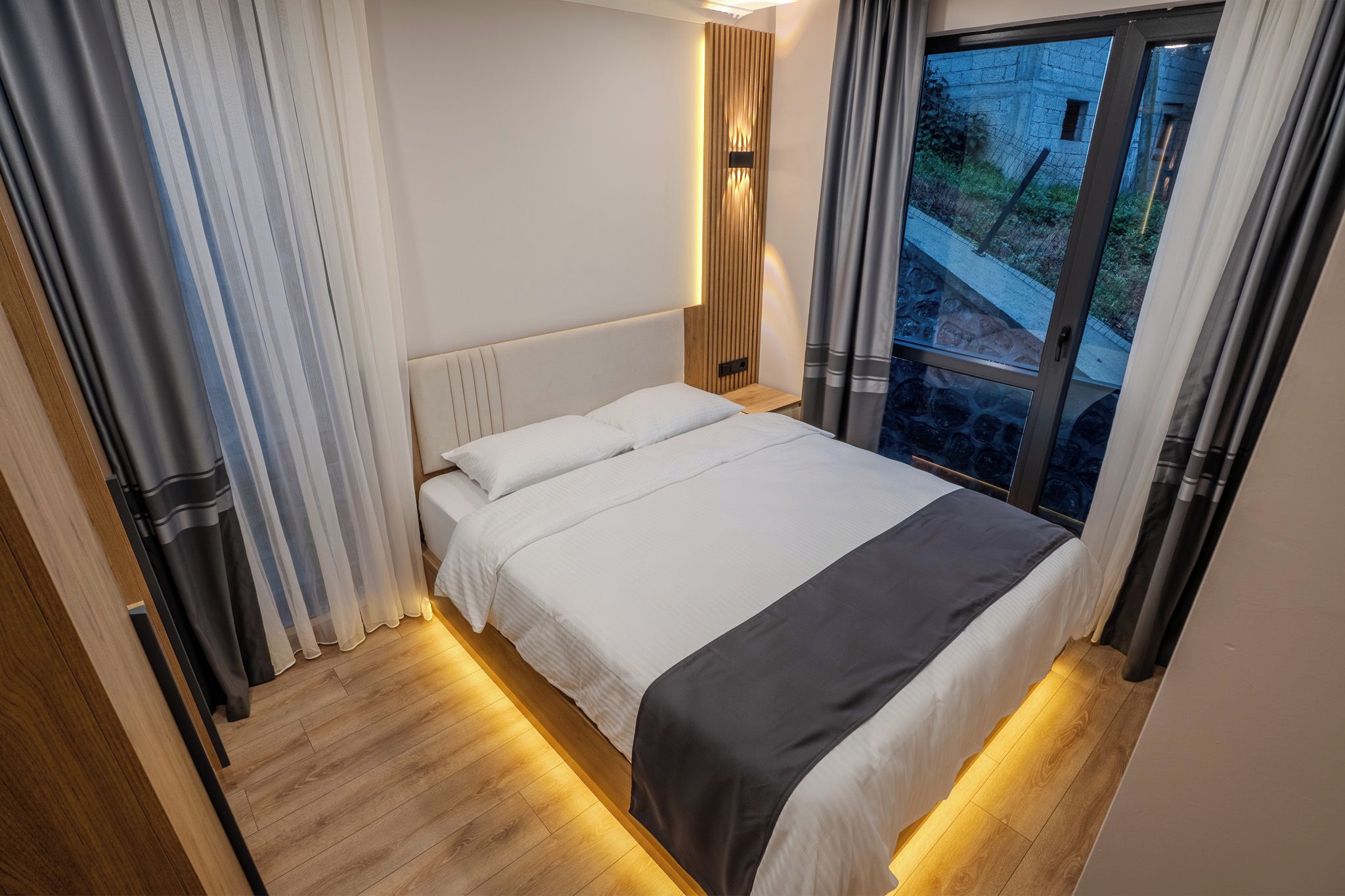 A modern bedroom with a large bed, wooden headboard, and a view of the outside through a window.