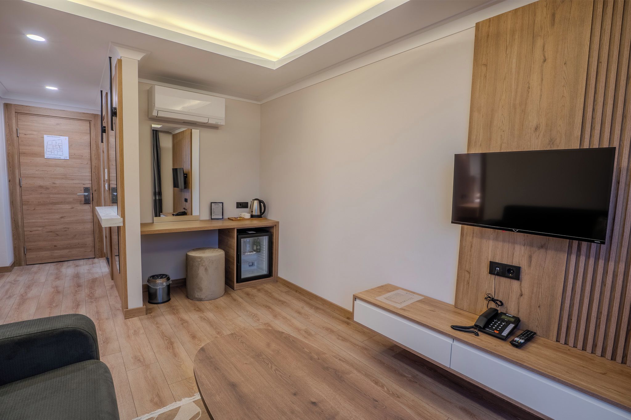 A hotel room with a wooden floor, a flat-screen TV mounted on the wall, a small refrigerator, a coffee maker, and a trash bin.