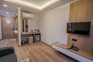 A hotel room with a wooden floor, a flat-screen TV mounted on the wall, a small refrigerator, a coffee maker, and a trash bin. - Zaya Residence (Yomra)