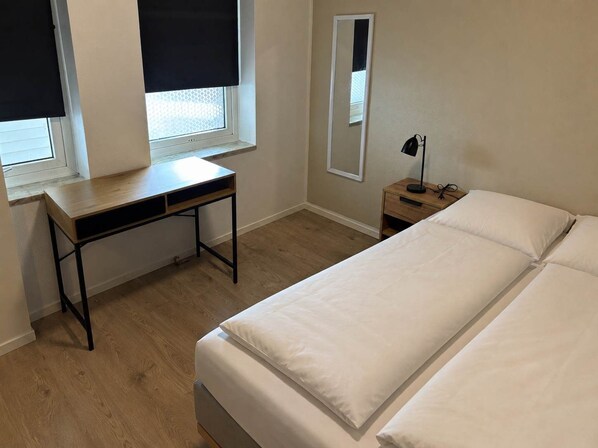 A bedroom with a bed, a desk, a nightstand, a mirror, and a window with blinds. - Klaksvik Mountain View Stay (Klaksvík)