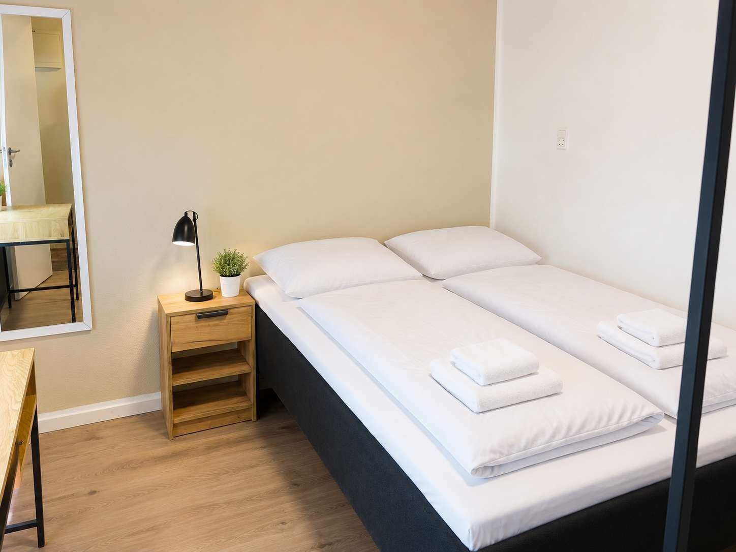 Ein Einzelbett mit weißer Bettwäsche und zwei Kissen, ein Nachttisch mit Lampe sowie ein kleiner Blumentopf.