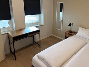 A bedroom with a bed, a desk, a nightstand, a lamp, and a window with blinds. - Klaksvik Mountain View Stay (Klaksvík)