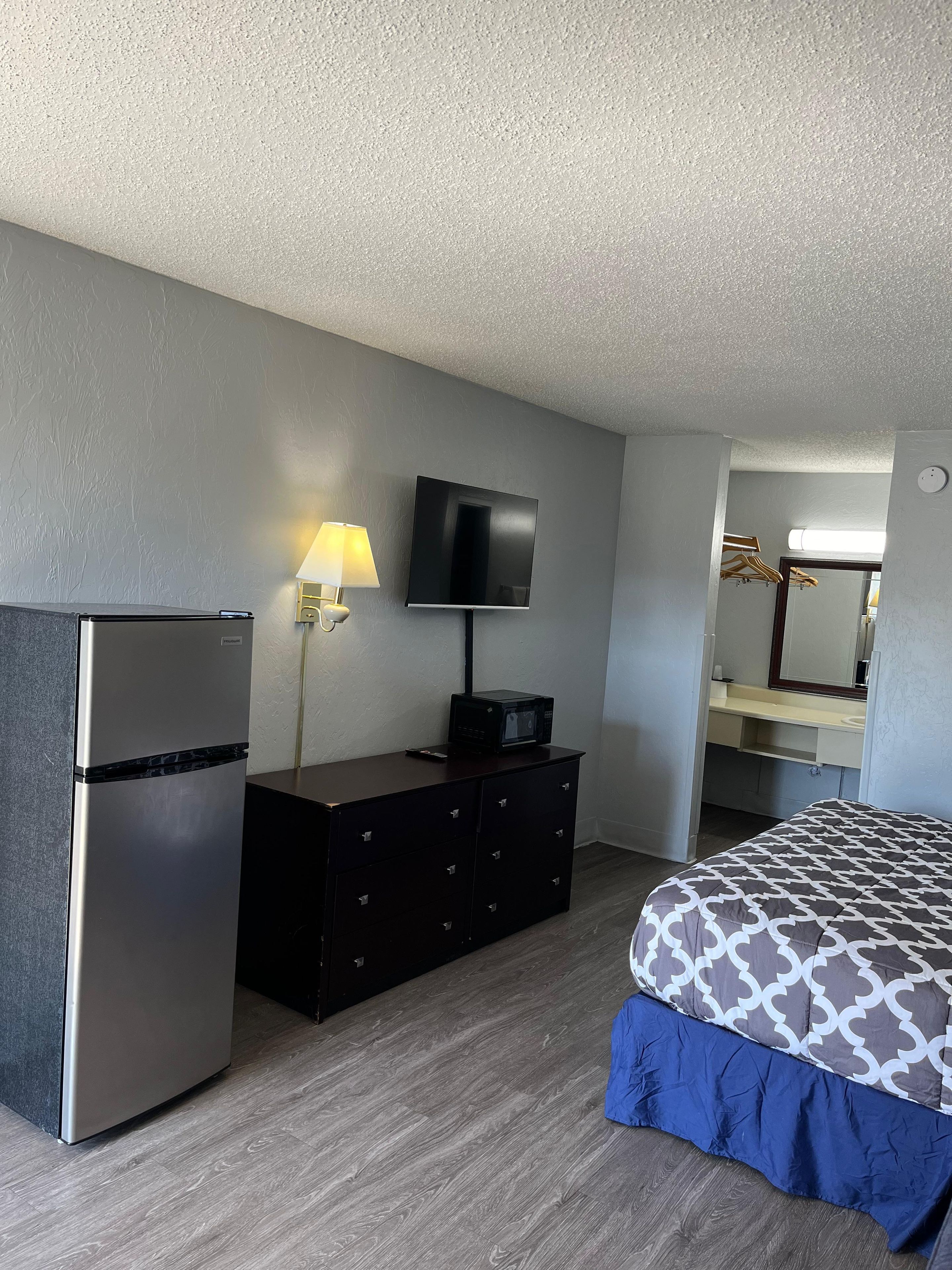 A hotel room with a bed, a dresser, a refrigerator, a flat-screen TV mounted on the wall, and a mirror above the dresser.