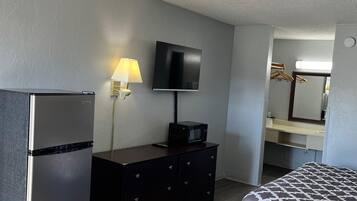 A hotel room with a bed, a dresser, a refrigerator, a flat-screen TV mounted on the wall, and a mirror above the dresser.