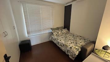 Una habitación pequeña y poco amoblada con una cama individual, un escritorio y una ventana con persianas.