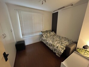 Une petite pièce, peu meublée, avec un lit simple, un bureau et une fenêtre avec des stores.