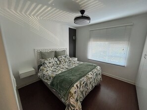 Ein Schlafzimmer mit einem Bett, einem Fenster mit Jalousien, einem Deckenventilator und einem Nachttisch.
