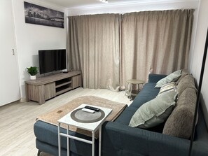 Ein Wohnzimmer mit einer Couch, einem Couchtisch, einem Fernseher auf einem Holzboden und einem Vorhang.