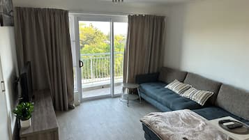 A modern living room with a sofa, a small side table, a television, and a balcony with curtains.