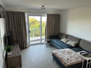 Ein modernes Wohnzimmer mit einer Couch, einem kleinen Beistelltisch, einem Fernseher und einem Balkon mit Vorhängen.