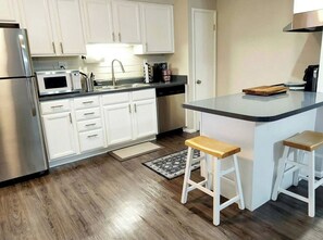 A kitchen with white cabinets, a stainless steel refrigerator, a microwave, and a coffee maker. There are two wooden stools and a small wooden cutting board on the counter.