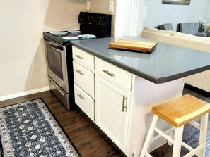 A kitchen with a stove, oven, and countertop. There is a wooden cutting board and a stool.