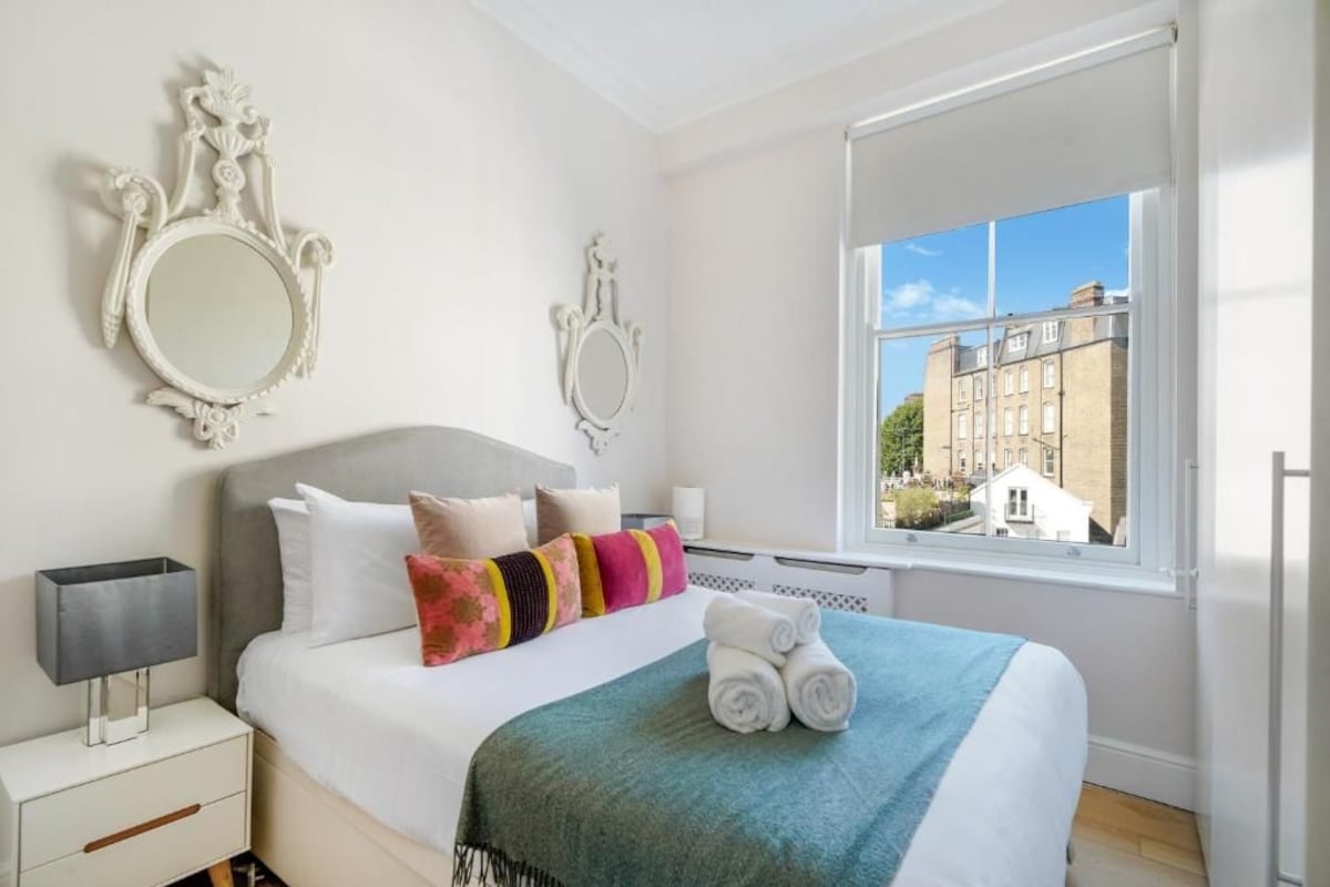 A hotel room with a bed, two wall-mounted mirrors, a window with a view of buildings, a bedside table with a lamp, and a rolled towel on the bed.