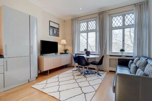 A modern living room with a sofa, a TV, a dining table with chairs, and a large window with curtains. -  Comfortable Apartments in Kensington by Short Stories London (London)