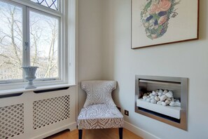 A corner room with a radiator, a window with a view of bare trees, a patterned chair, and a fireplace with white stones. -  Comfortable Apartments in Kensington by Short Stories London (London)
