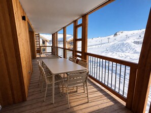 Eine Holzdielen-Terrasse mit Tisch und Stühlen, mit Blick auf eine verschneite Berglandschaft und Skilifte.