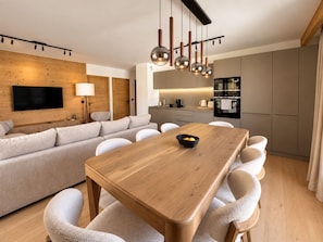 Ein modernes Wohnzimmer mit einer Couch, einem Esstisch mit Stühlen, einem an der Wand befestigten Fernseher und einer Küchenzeile mit Schränken und Geräten.