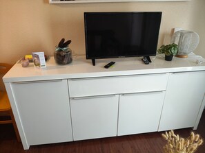 A white entertainment center with a flat-screen TV, remote controls, a candle, a decorative glass jar, and a potted plant. - Bright apartment - 4 guests south view (Besse-et-Saint-Anastaise-Super Besse)
