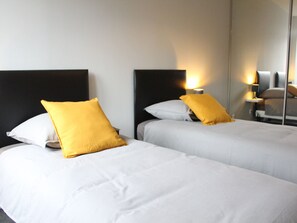 Une chambre d’hôtel avec deux lits, chacun doté de draps blancs et d’un seul oreiller jaune. Des lampes de chevet sont placées de chaque côté des lits.
