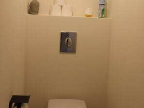 Une salle de bain avec des toilettes, une étagère contenant des articles de toilette et un interrupteur fixé au mur.