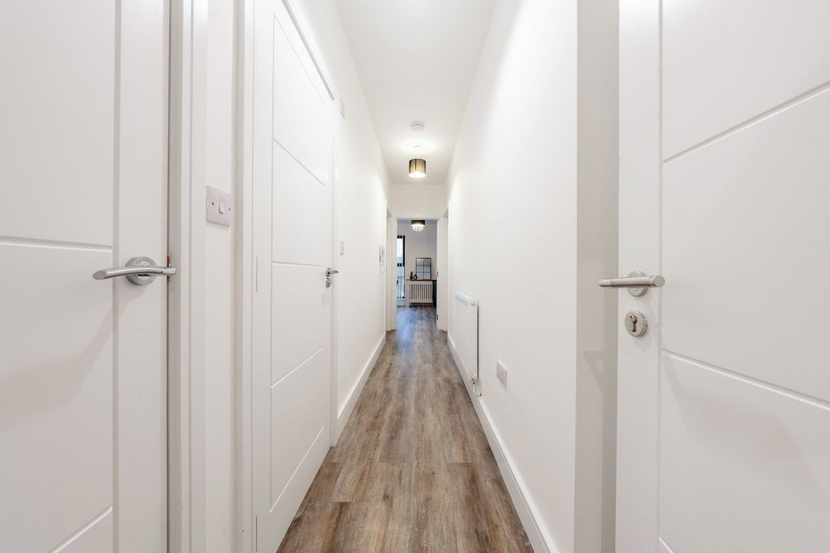 A narrow hallway with white doors and a wooden floor, leading to a room with a radiator and a chandelier.