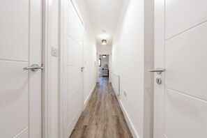 A narrow hallway with white doors and a wooden floor, leading to a room with a radiator and a chandelier. - Sleeps 4 | 2 Bed | Free Parking | Potters Bar (Potters Bar)