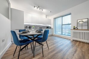 A modern kitchen with white cabinets, a dining table with blue chairs, and a large window with blinds. - Sleeps 4 | 2 Bed | Free Parking | Potters Bar (Potters Bar)