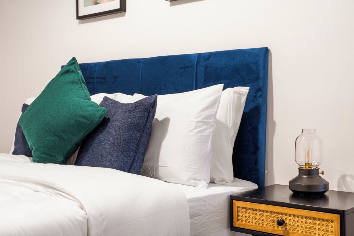 A bed with a blue headboard, white and green pillows, and a bedside table with a lamp.