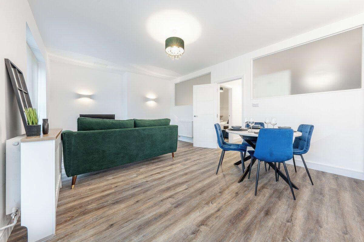 A modern living room with a green sofa, a dining area with blue chairs, and a wooden floor.