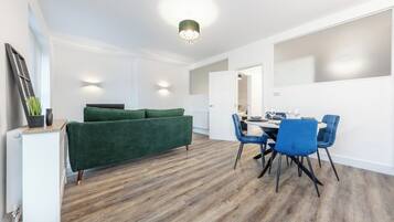 A modern living room with a green sofa, a dining area with blue chairs, and a wooden floor.